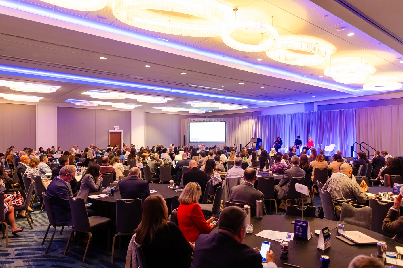 Wide view of a full conference ballroom with a glowing stage and ceiling lights.