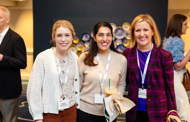 Three attendees smiling together with lanyards in front of a wall of decorative plates.