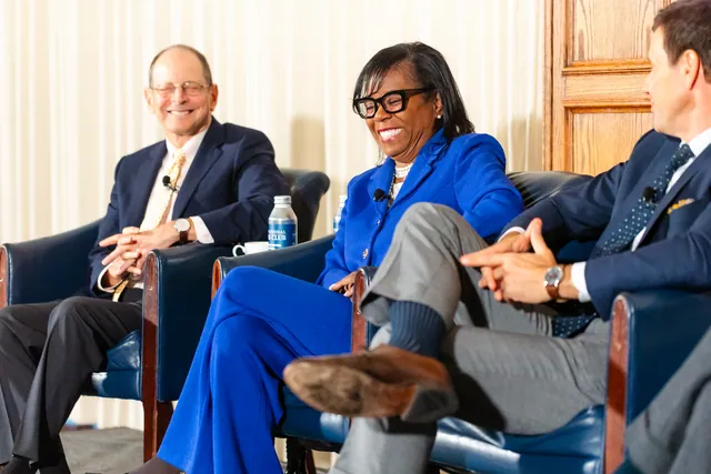 Panel guests laugh together on stage in blue armchairs.