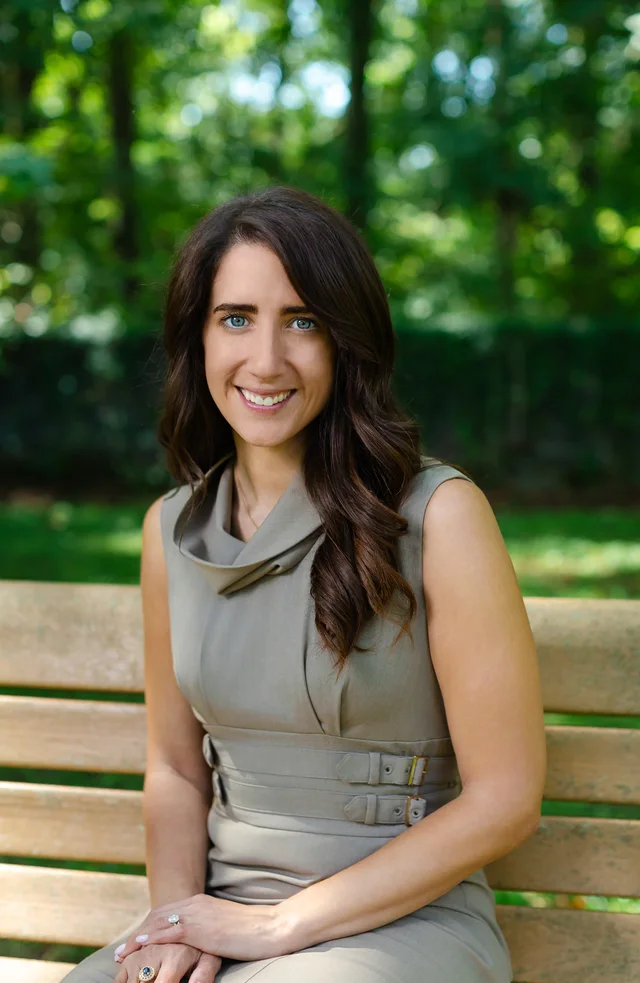 A professional seated on a park bench, smiling warmly at the camera in a gray sheath dress, with trees behind her.
