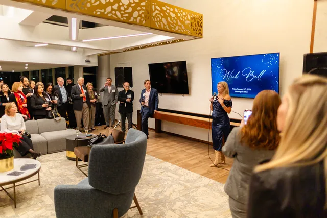 Guests gather in a bright lounge to listen to a speaker beside an event title screen.