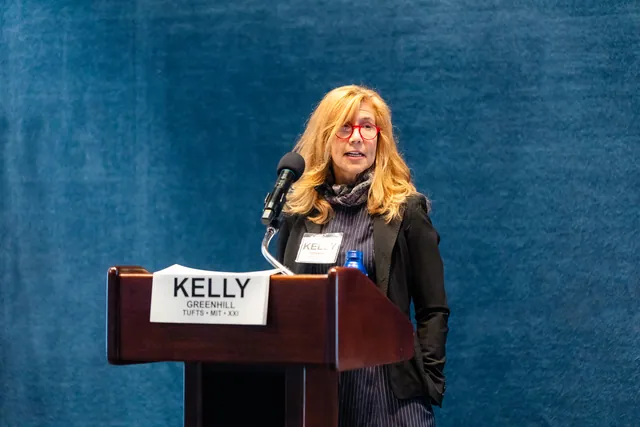 Speaker with long blonde hair addresses the room from a podium with a name card.
