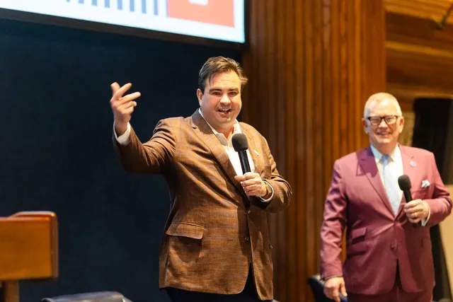 Presenter mid-gesture with a microphone beside a co-host on a wood-paneled stage.