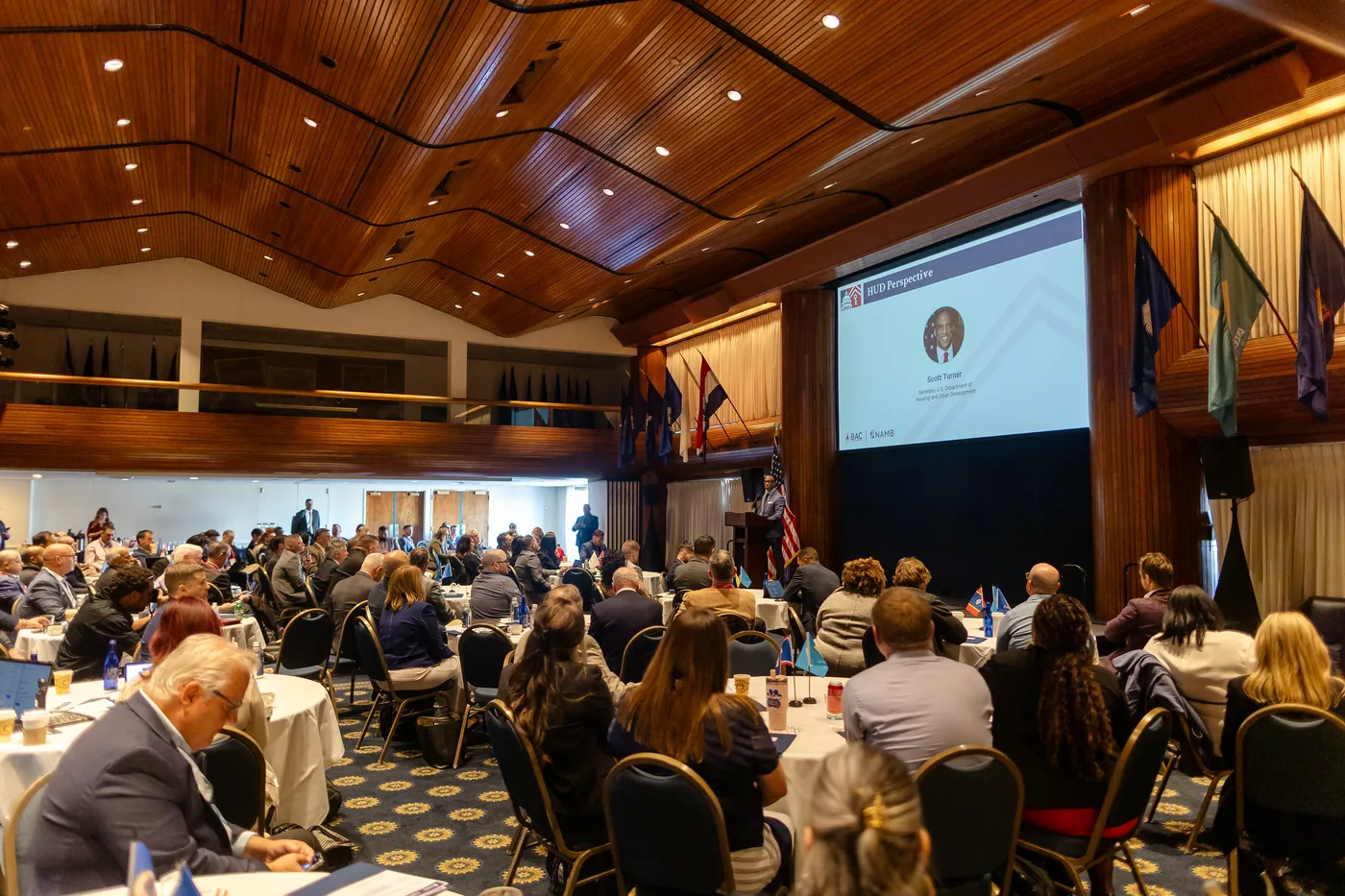 Wide view of a packed ballroom with wood ceiling and audience seated at round tables facing a screen.