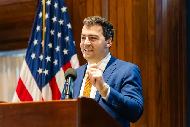 Speaker in a blue suit at the podium smiling mid-gesture in front of an American flag.
