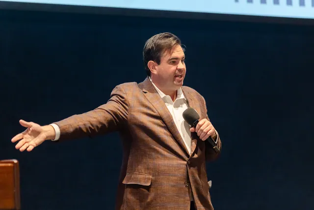 Presenter in a brown blazer with arms stretched wide while speaking into a handheld microphone.
