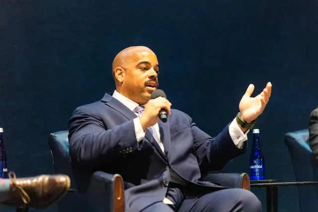 Panelist in a navy suit gesturing with open hands while speaking on stage.