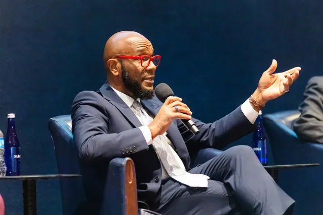 Panelist in red-framed glasses gesturing broadly while speaking into a microphone.