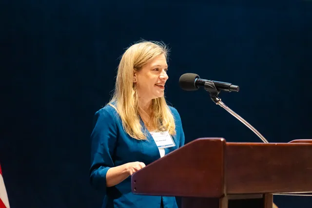 Speaker in a blue blazer speaking at a wooden podium against a deep navy curtain.