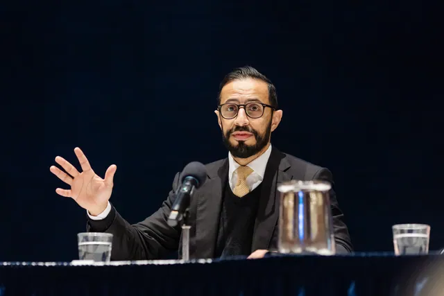 Panelist with glasses gestures with an open hand while speaking at a draped table.