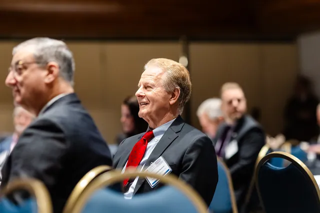 Attendee in a red tie smiles broadly while listening in the audience.