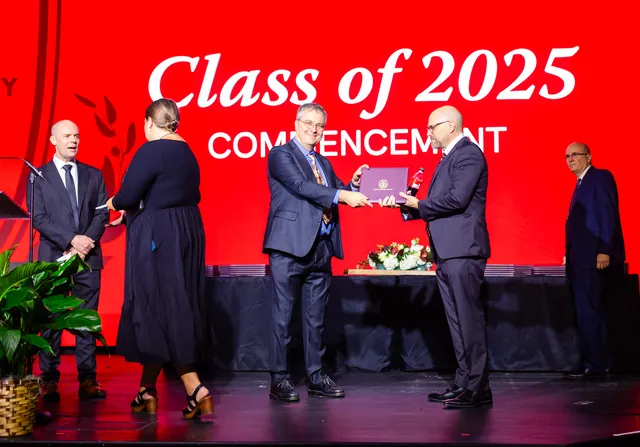 A graduate receives a diploma and handshake on stage in front of a large red commencement screen.