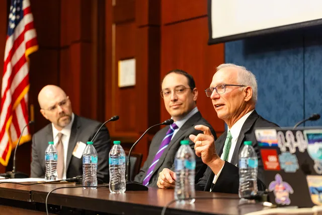 Panelists speak and listen at a long table with water bottles and an American flag behind.