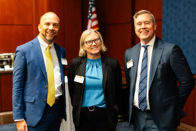 Three attendees smile together at a reception in the wood-paneled room.