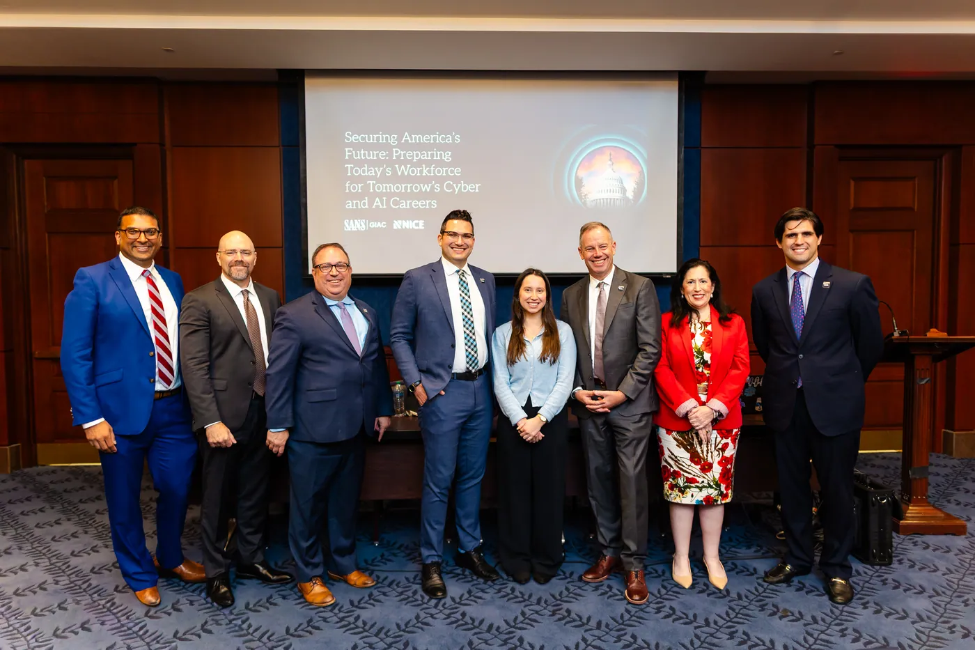 A group of eight speakers and guests pose together in front of the presentation screen.