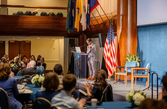 Speaker at lectern with flag, audience in foreground, balcony and flags behind.