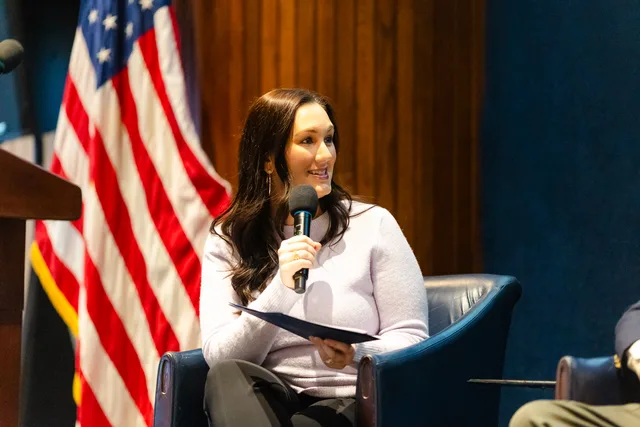 Moderator speaking with flag behind her, deep navy background.
