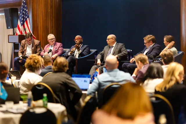 Panel under the flag, seen over the heads of an attentive audience.