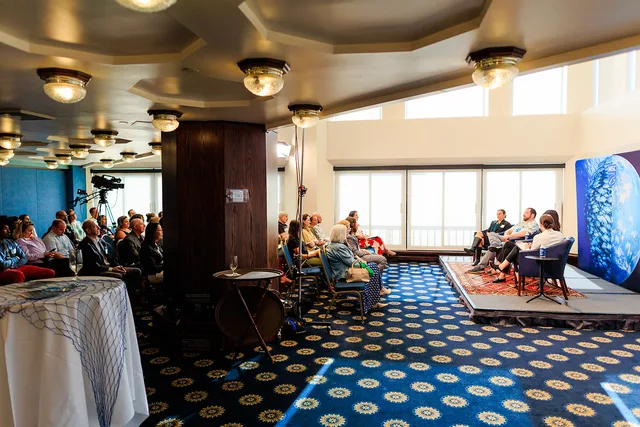 Wide view of a bright room with a panel seated on a rug-covered platform and the audience beside it.