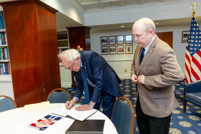 Bernie Sanders signing a photograph beside a National Press Club staff member, flags behind them.