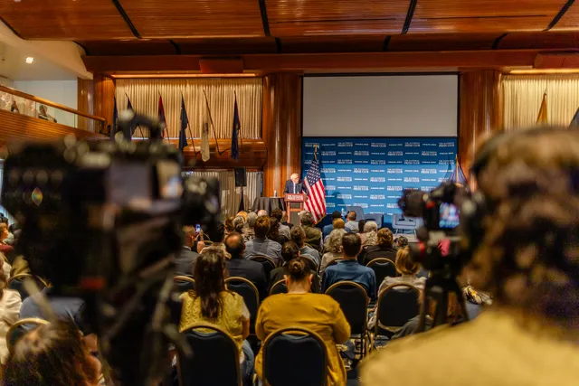Press cameras in the foreground frame a packed National Press Club hall as Bernie Sanders speaks from the podium far ahead.
