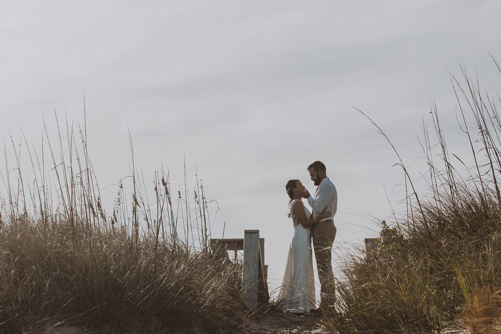 A couple standing forehead to forehead in the dunes, framed by sea oats against a pale sky