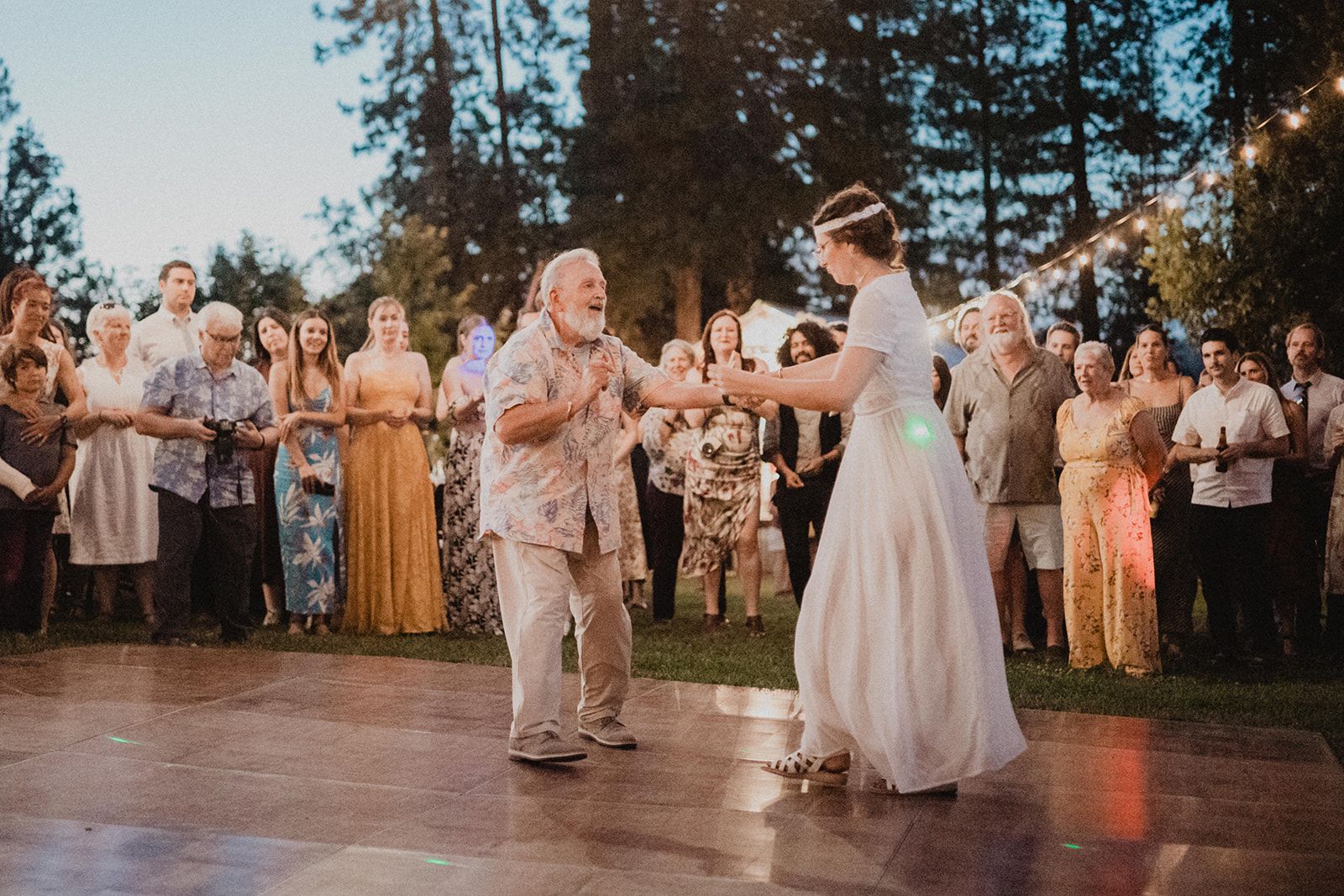 An older man in a patterned shirt dances with the bride on an outdoor dance floor while guests watch from the edge