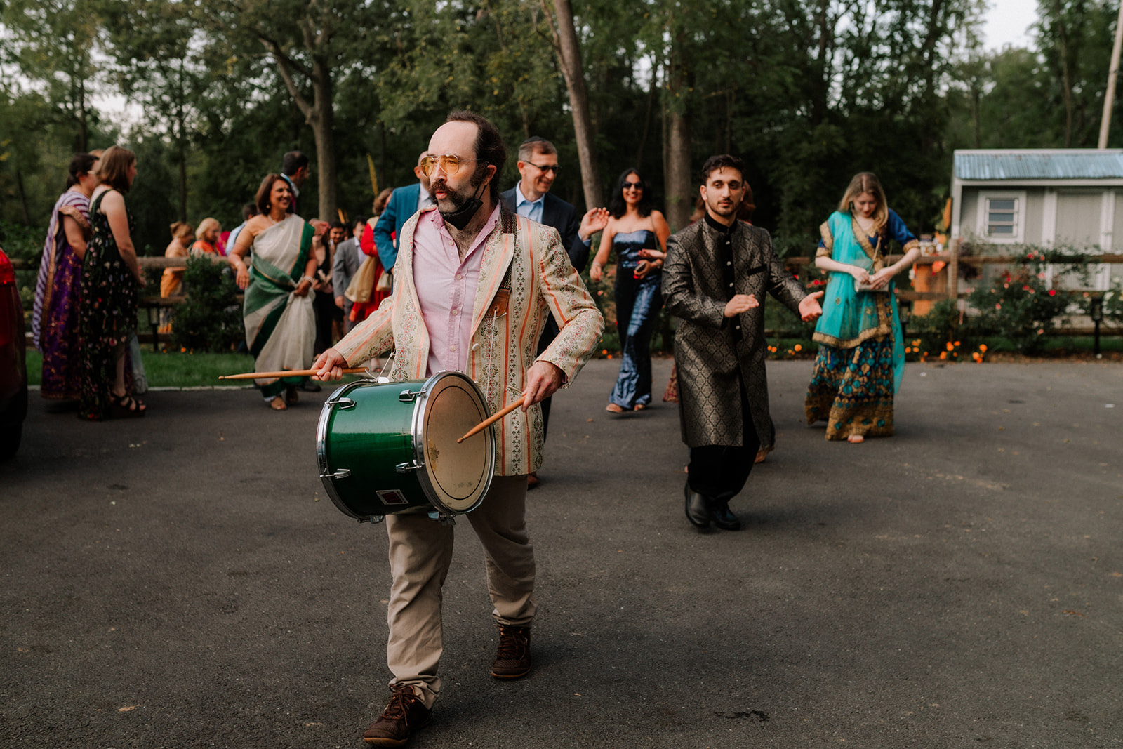 A drummer leading the baraat processional down the driveway, guests in sherwanis and sarees following