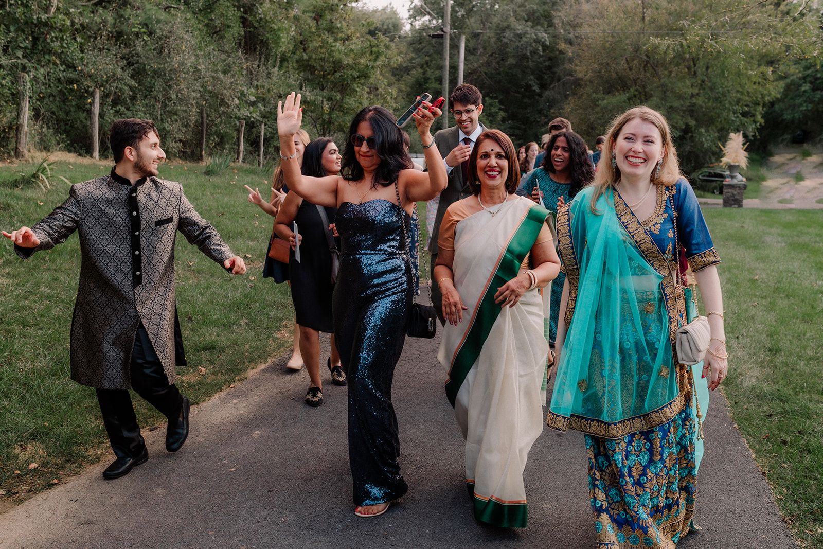 A group of guests in saris and formal wear walks along a paved path together, arms up and faces grinning, one raising a phone above the crowd