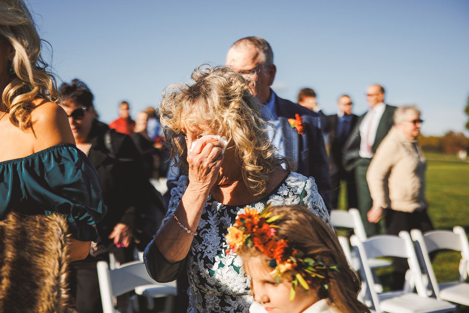 An older woman dabs her eye with a tissue among the white ceremony chairs while a girl in a bright flower crown stands in front of her, head lowered