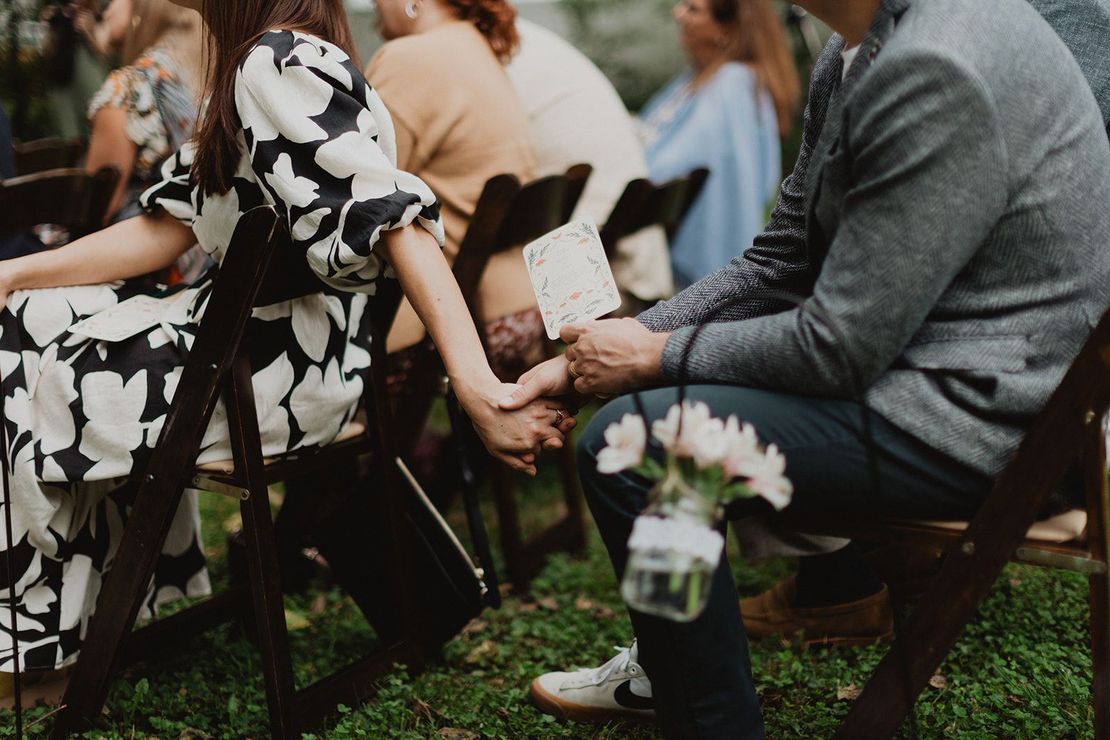 Two guests seated in ceremony chairs hold hands while one of them holds a small printed card
