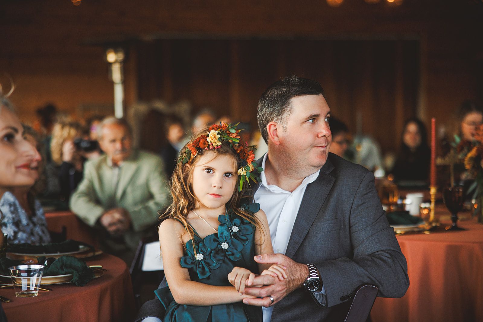 A young girl in a flower crown leans back against a man in a grey suit at a reception, her hands folded into his