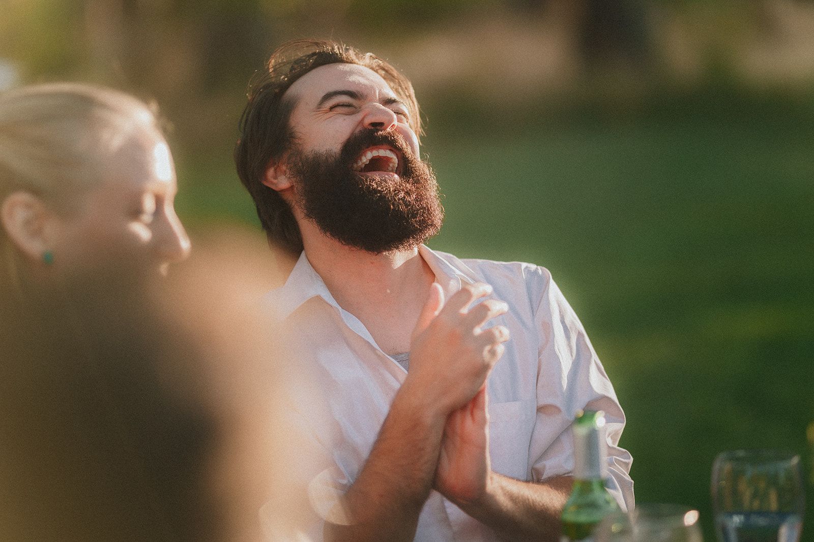A bearded man in a white shirt tips his head back mid-laugh with his palms pressed together at his chest, an out-of-focus guest in the foreground