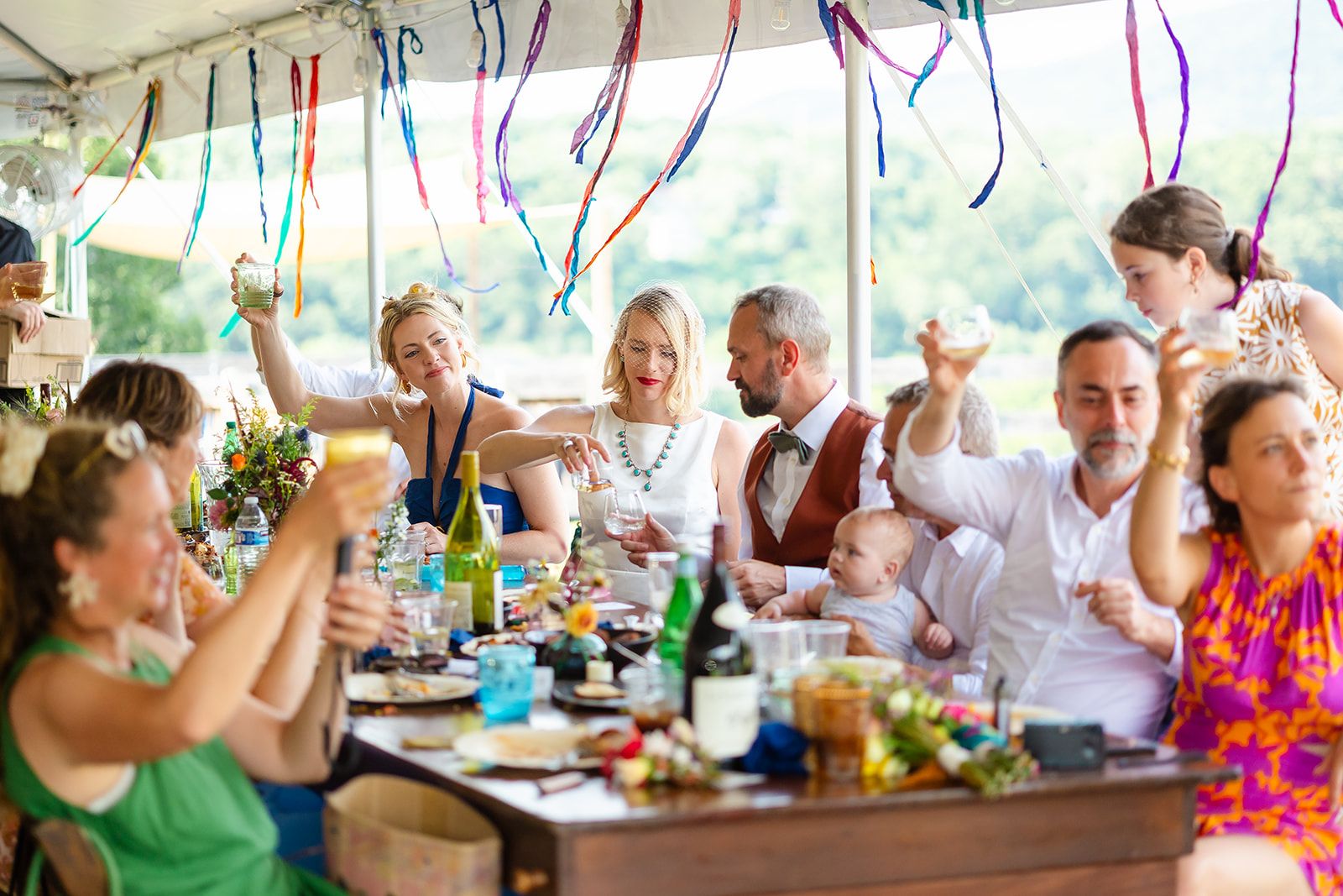 Guests along a long table under a streamer-hung tent raise their glasses in a toast while a baby sits on a lap in the middle of the row