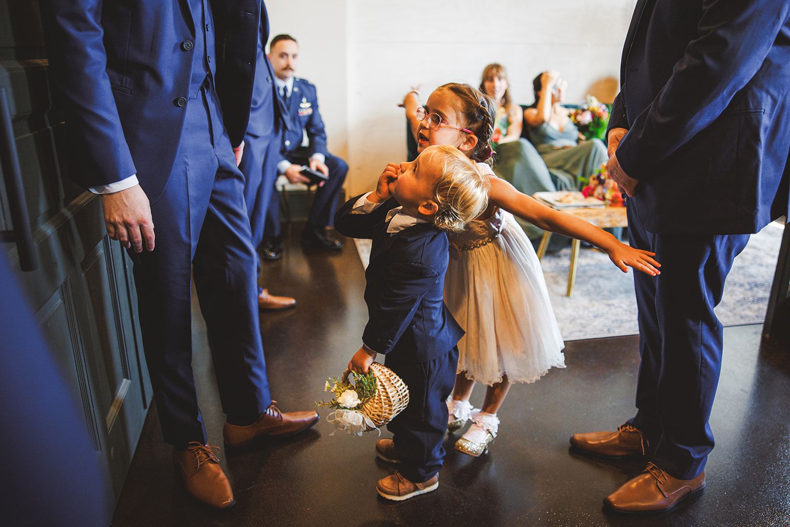Two small children stand pressed together between grown-ups in navy suits, the girl in a tulle dress with her mouth wide open and one arm flung out while the boy in a navy suit leans back into her holding a flower basket.
