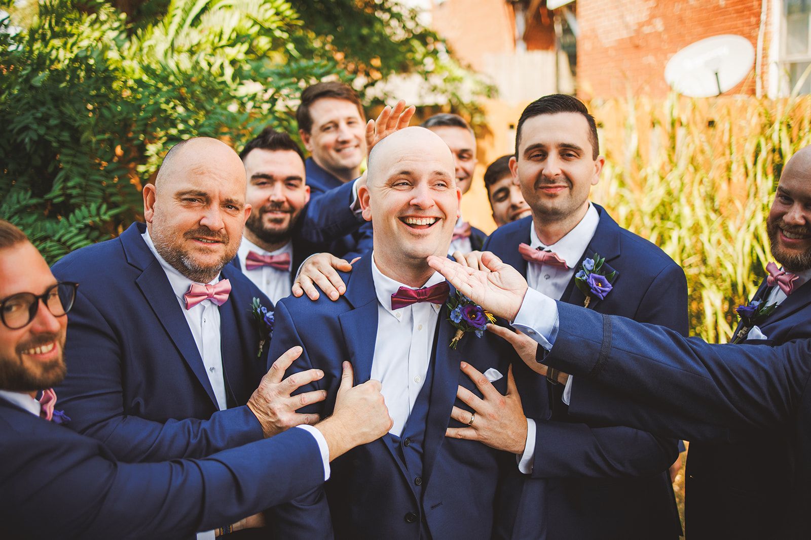 Groomsmen in navy suits and dusty pink bow ties crowd in and press their hands to one laughing man's chest and shoulders.