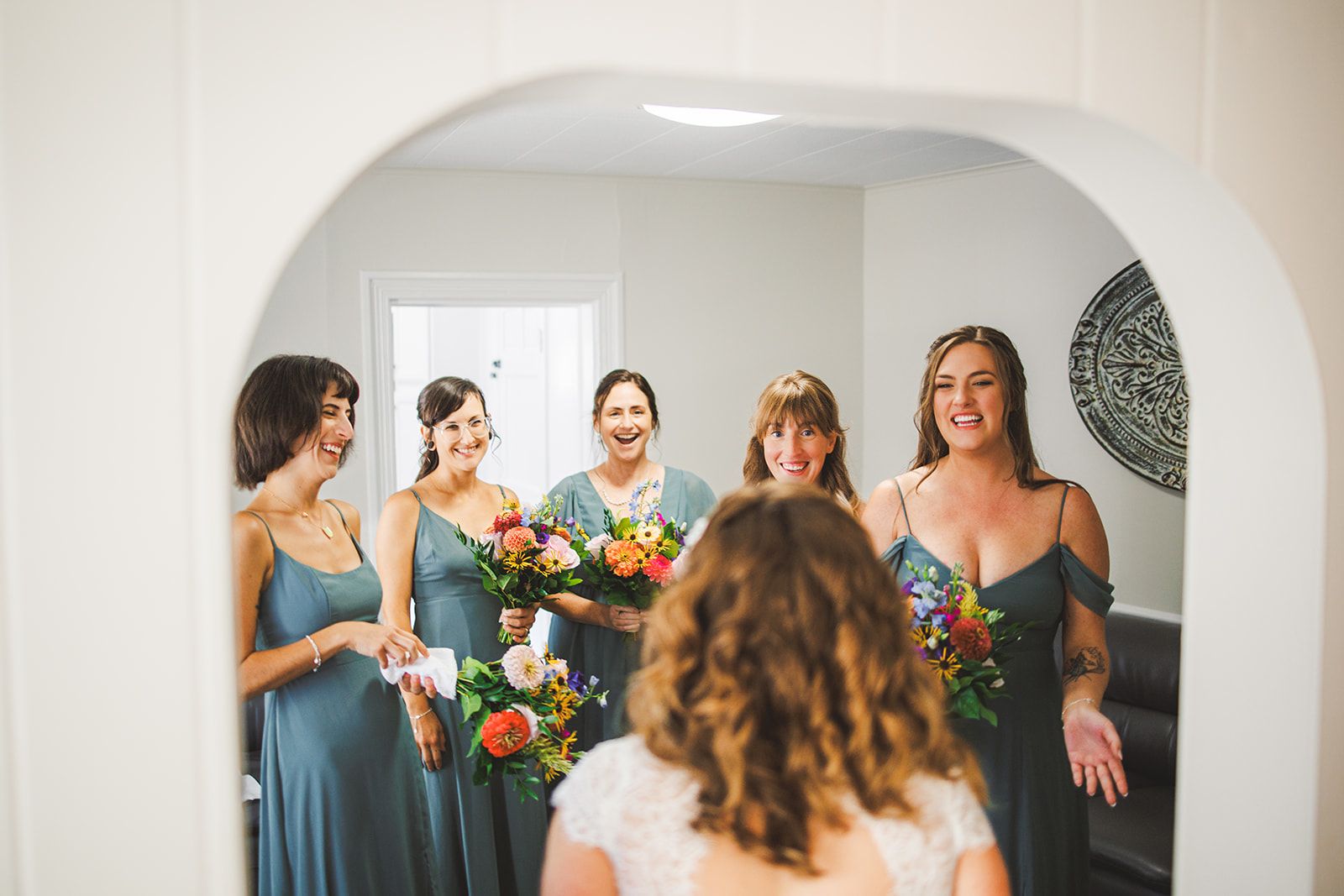 Five women in matching sage green dresses light up laughing through an archway as a woman in a white lace dress steps in front of them with her back to the camera.