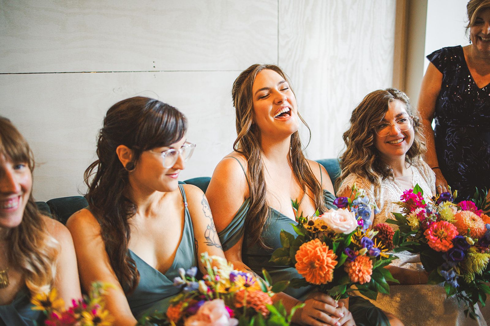 Women in sage green dresses sit shoulder to shoulder with bright bouquets in their laps, one of them laughing with her eyes closed and head tipped back.