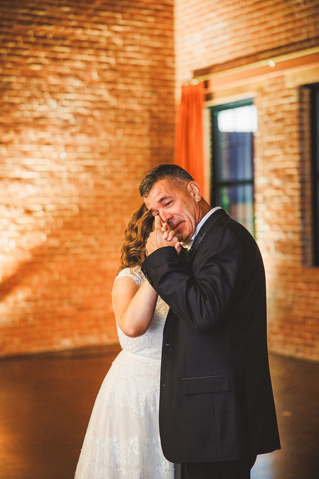 An older man in a dark suit holds a woman in a white lace gown close in an empty brick room and wipes his eye, his face crumpling.