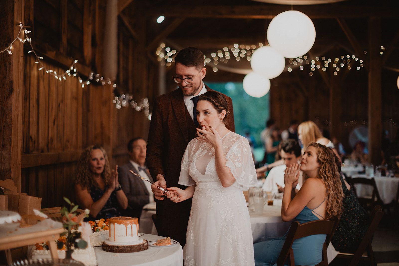 The bride tastes cake off her fingertips at the cake table while the groom smiles down at her and seated guests clap.