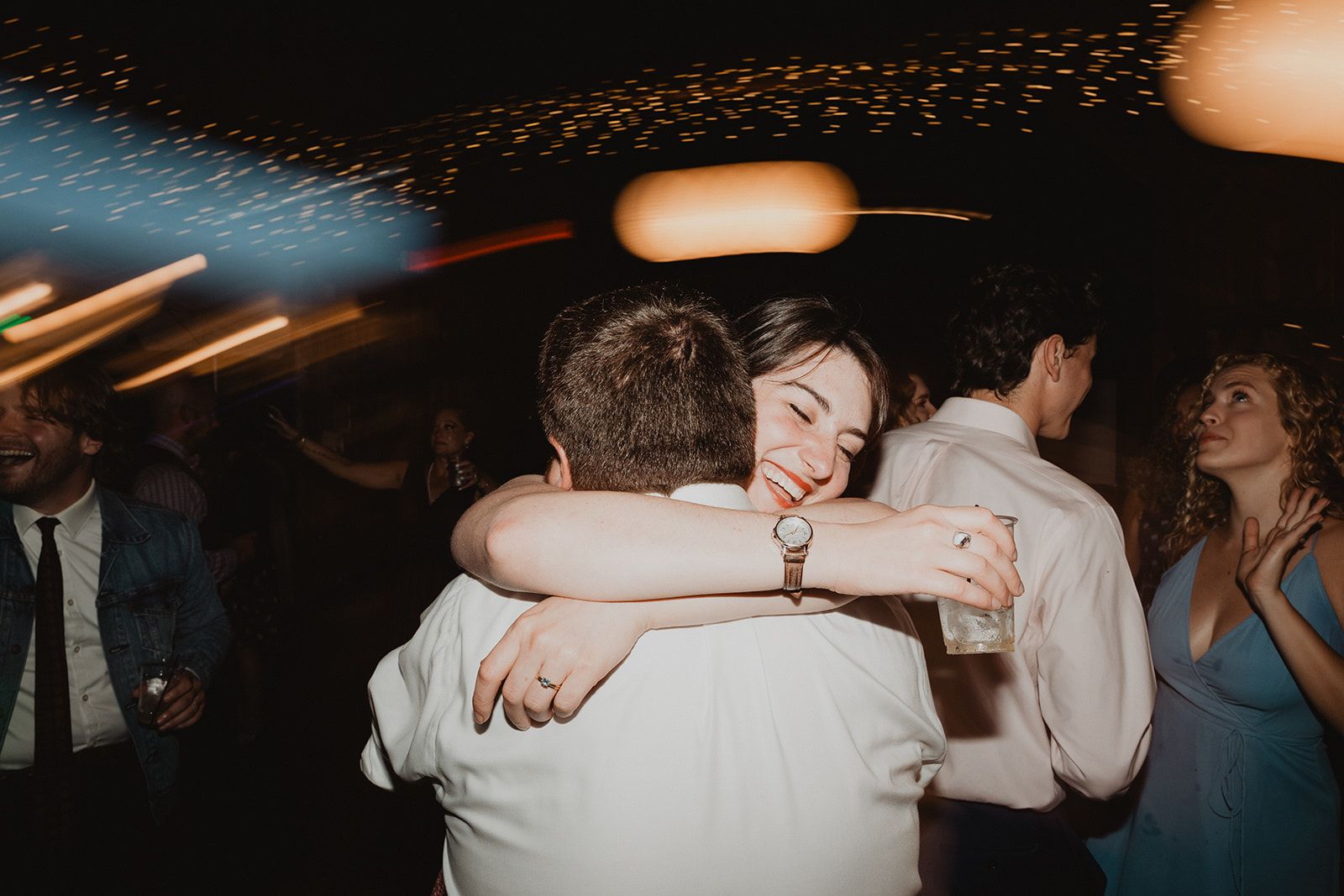 A woman throws both arms around a man's neck in a tight hug at the reception, still holding her drink, eyes closed and smiling.