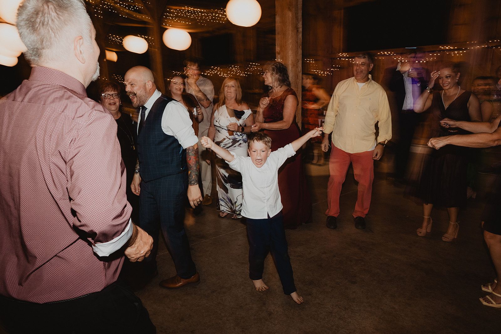 A barefoot young boy dances with both arms flung wide while adults dance around him on the reception floor.
