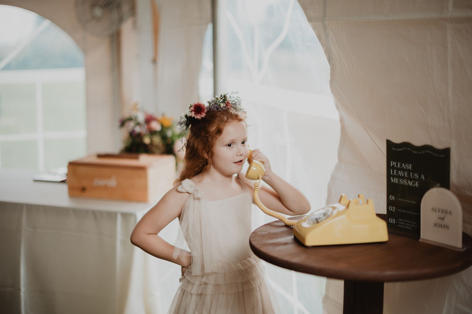 A young girl in a flower crown holds the handset of a yellow rotary phone to her ear with her other hand on her hip.