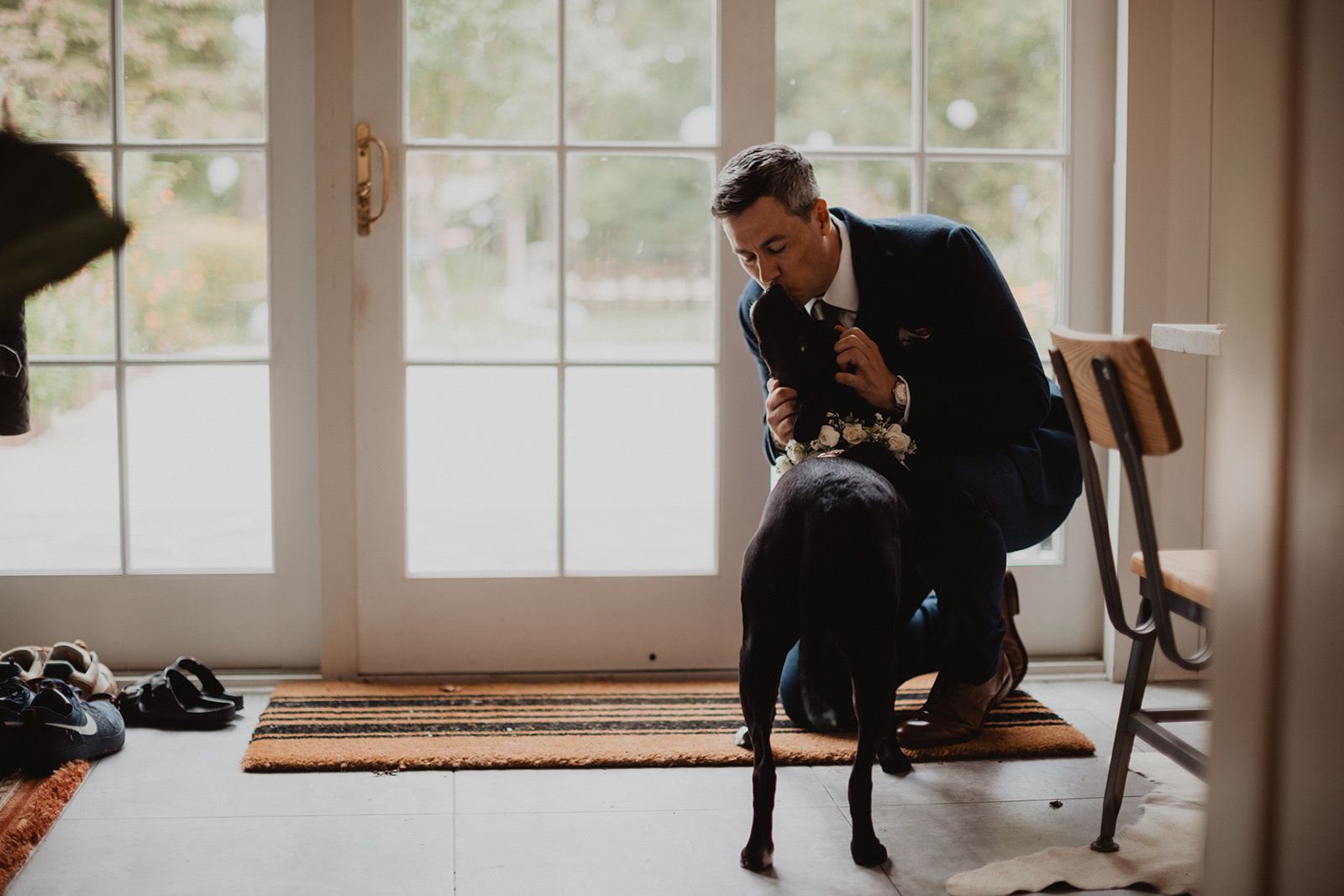 A man in a navy suit crouches down and leans his face close to a black dog wearing a flower collar.