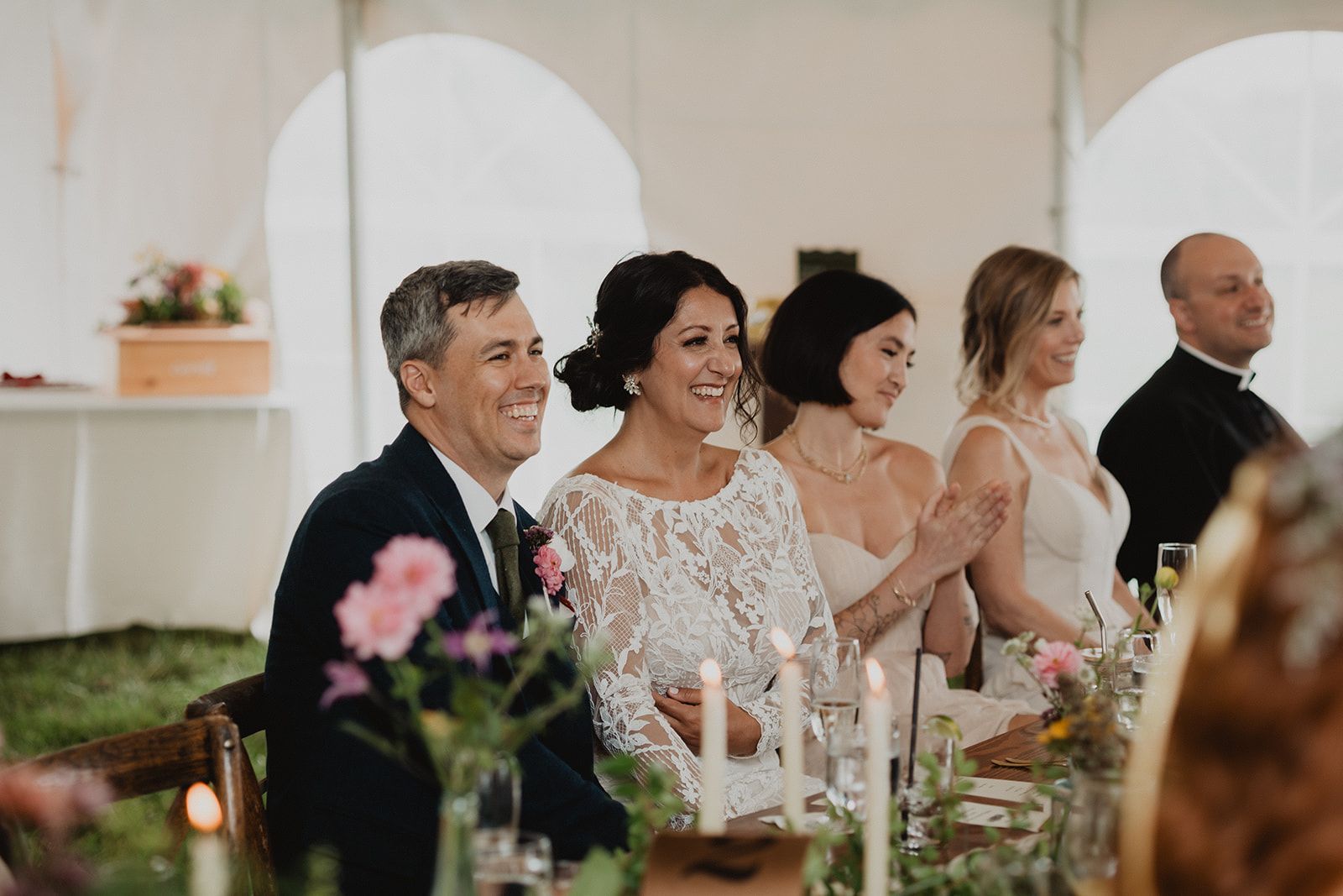 A row of guests at a long candlelit reception table smile and laugh as they watch something off camera, one of them clapping.