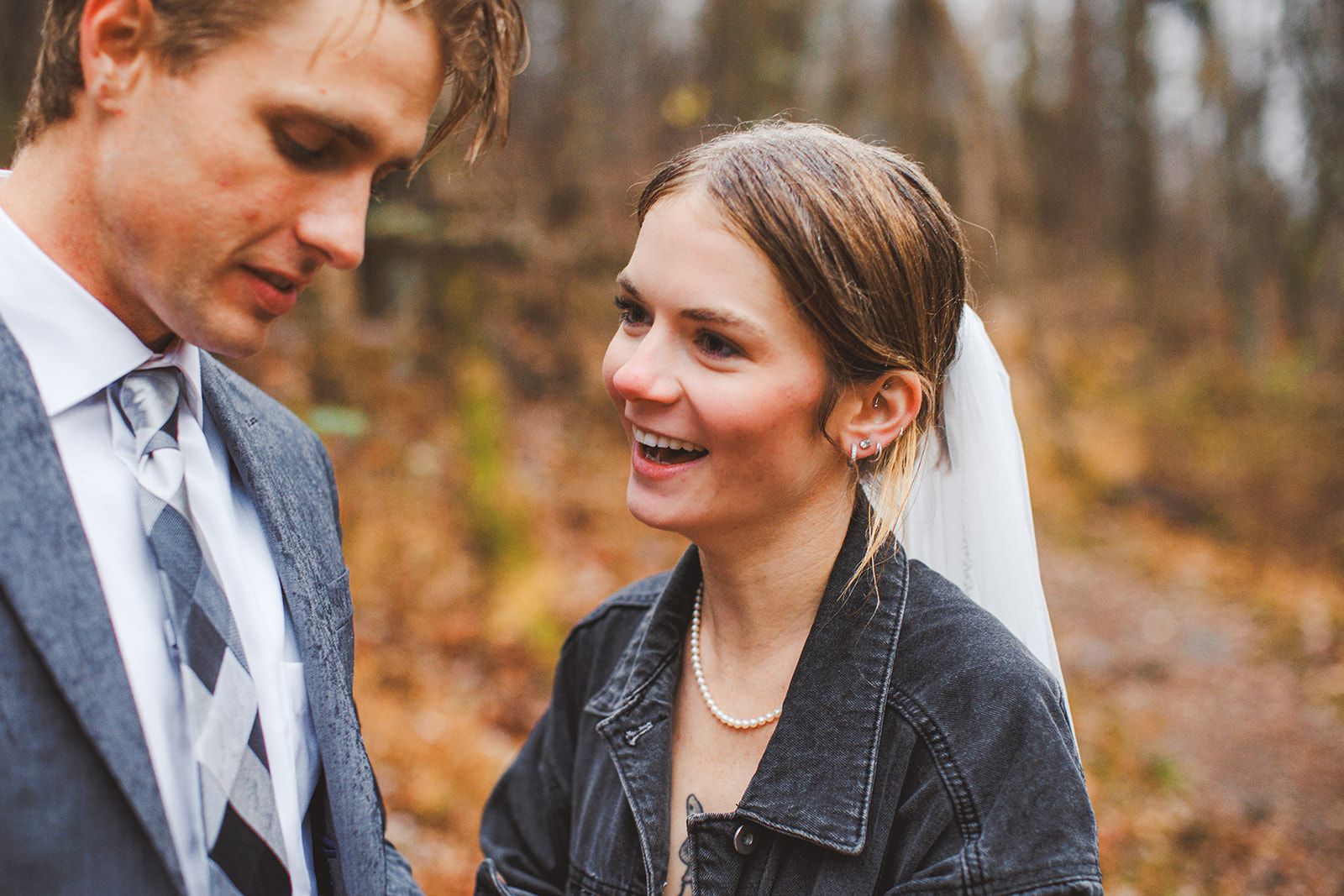 A woman in a dark denim jacket with her veil behind her laughs while looking off to one side as the man in a grey suit beside her lowers his head, blurred autumn woods behind them.