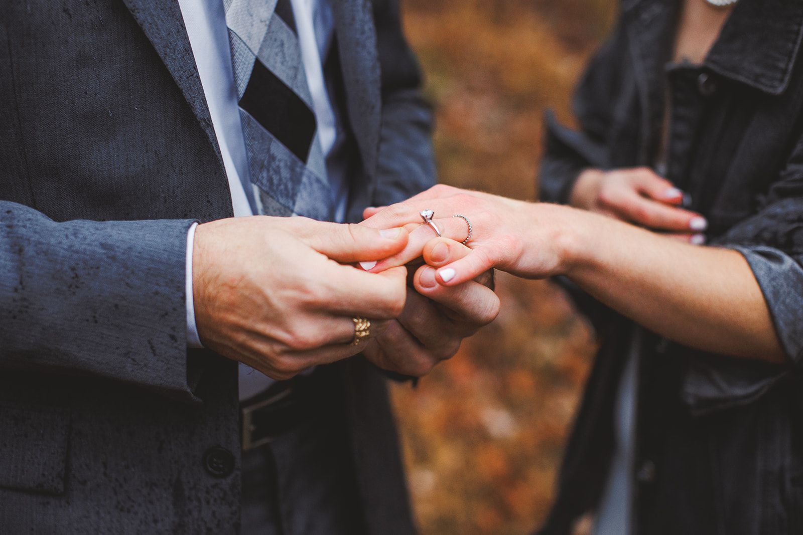 A man in a dark suit cradles a woman's hand in both of his, the rings on her finger catching the light against blurred autumn woods.