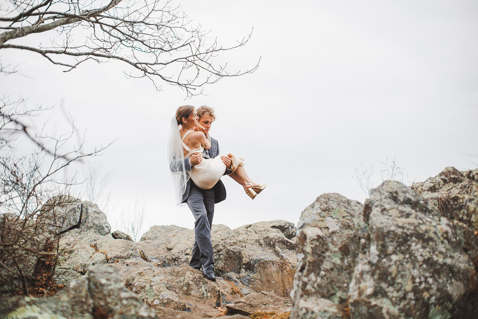 A groom in a grey suit carries the bride in his arms across a rocky outcrop, her long veil trailing and her arm wrapped around his shoulders.