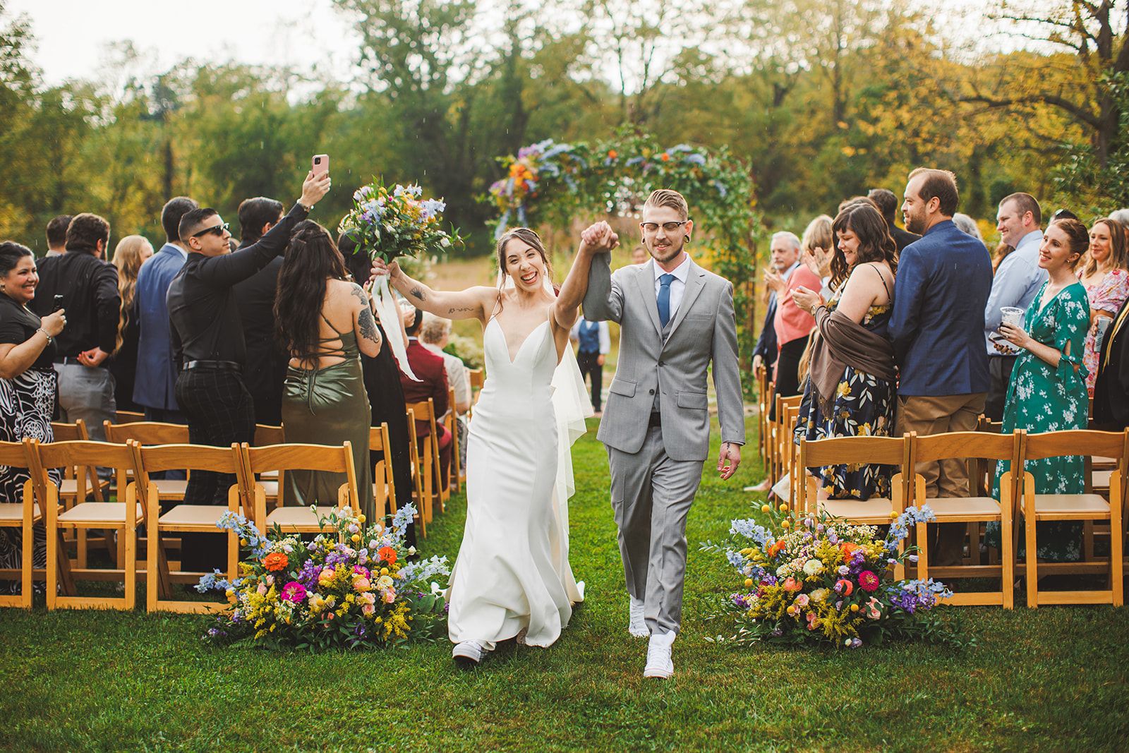 The newlyweds come back up the grass aisle with their joined hands and the bride's bouquet raised high while guests stand and applaud on both sides.