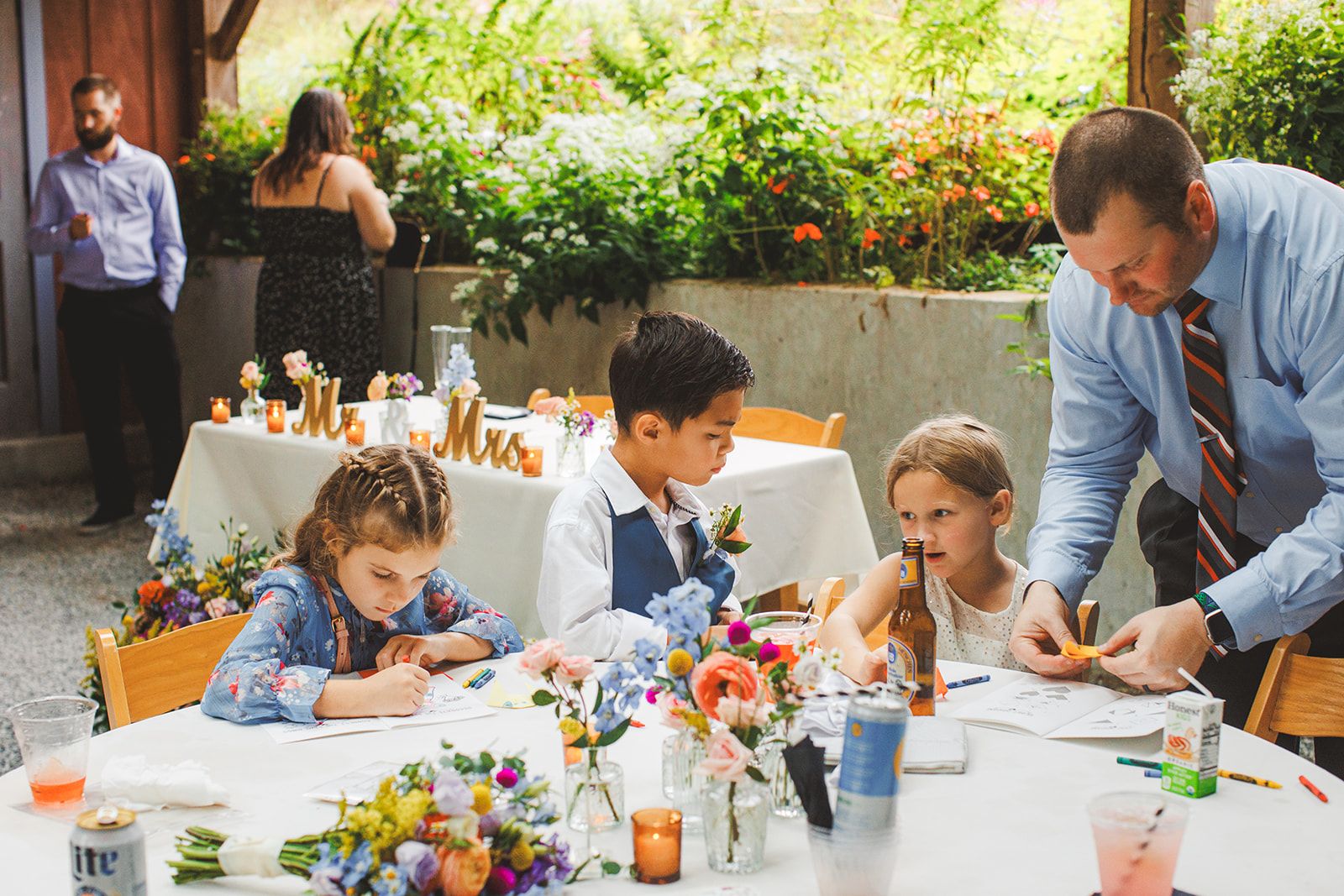 Three children work at a reception table spread with coloring pages and crayons while a man in a shirt and tie leans across to help the girl on the right with something in her hands.